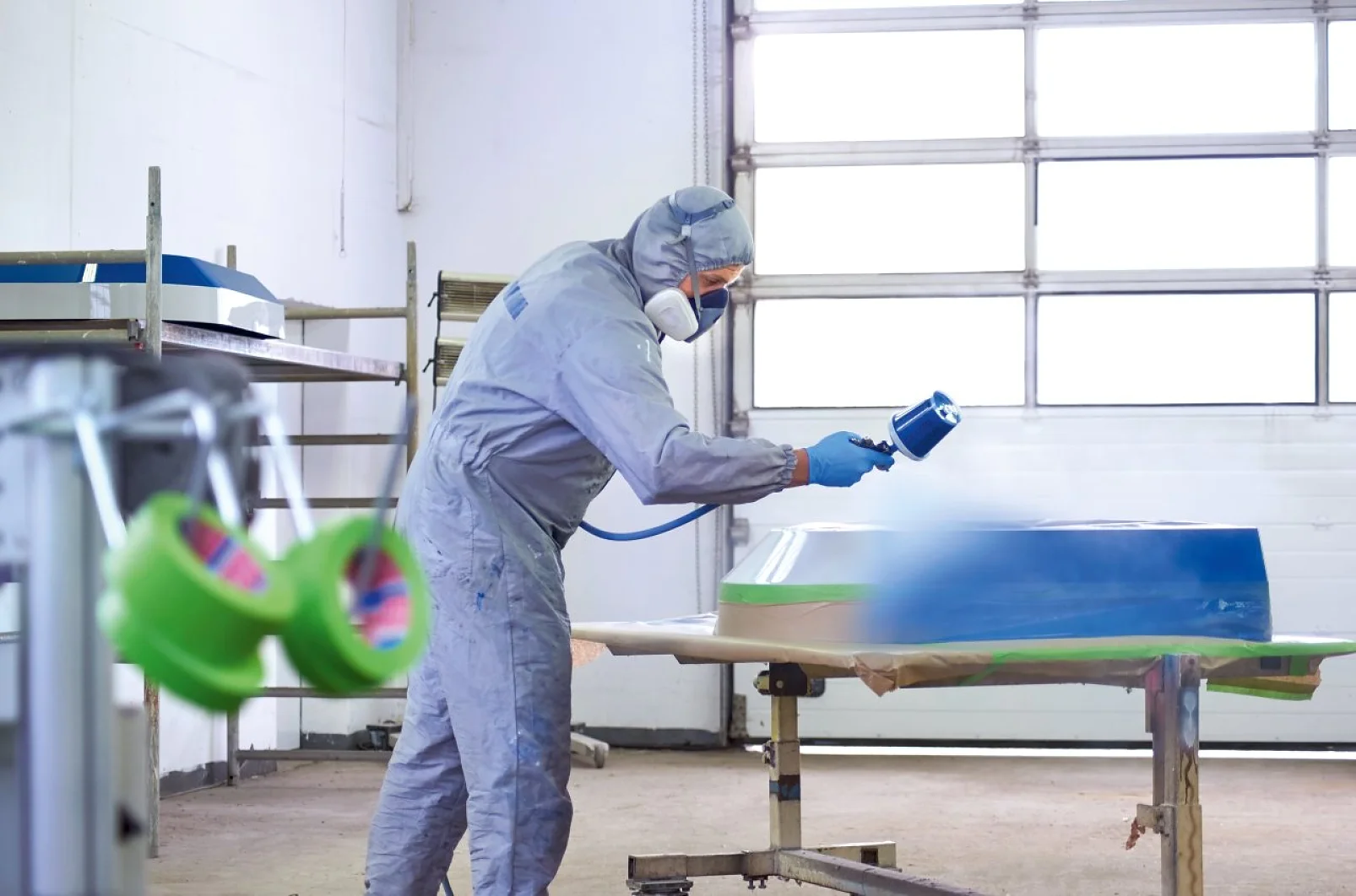 Eine Person in Schutzkleidung, darunter Maske und Overall, besprüht ein Objekt auf einem Tisch in einer Werkstatt. Der Bereich wirkt industriell, mit großen Türen und Regalen voller Materialien wie Tesa-Klebeband. (This text has been generated by AI)