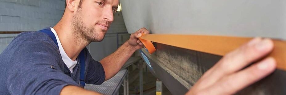 Ein Mann in blauem Hemd und Schürze klebt vorsichtig einen Streifen Tesa-Klebeband an der Kante einer großen, glatten Oberfläche entlang, möglicherweise dem Rumpf eines Bootes. Es scheint sich um eine Werkstatt zu handeln. Im Hintergrund ist eine Neonröhre zu sehen. (This text has been generated by AI)