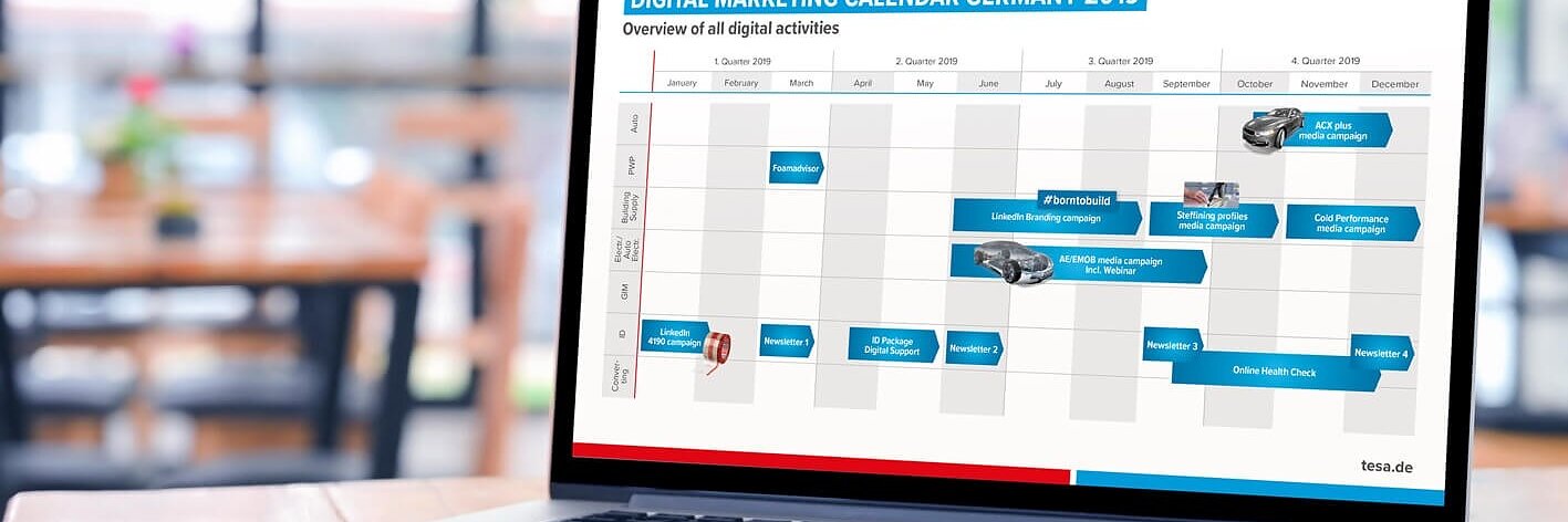 Auf einem Laptop auf einem Holztisch ist ein digitaler Marketingkalender für Deutschland 2019 von tesa zu sehen. Der Bildschirm zeigt ein Diagramm mit Monaten als Spalten und verschiedenen digitalen Aktivitäten, die über das Jahr verteilt geplant sind. Zur Planung und Organisation wird tesa-Klebeband verwendet. (This text has been generated by AI)