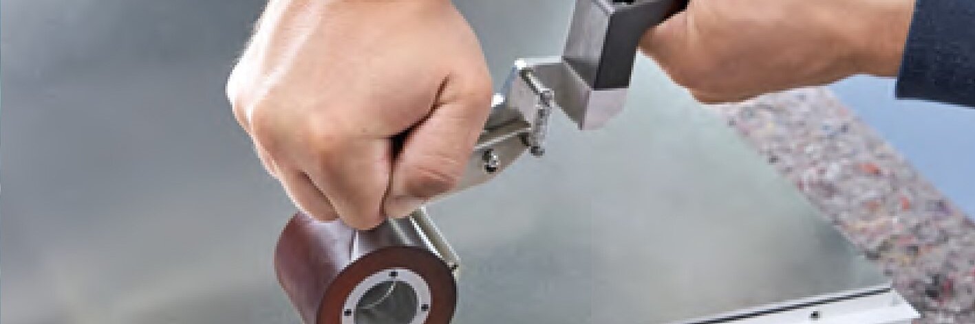 Eine Person verwendet ein Walzenwerkzeug auf einer flachen Glas- oder Metalloberfläche. Die Walze hat eine zylindrische Form und ist an einem Griff befestigt, der mit beiden Händen gehalten wird. Der Hintergrund umfasst Teile eines Stoff- und Metallrahmens, in einigen Abschnitten ist Tesa-Band sichtbar. (This text has been generated by AI)