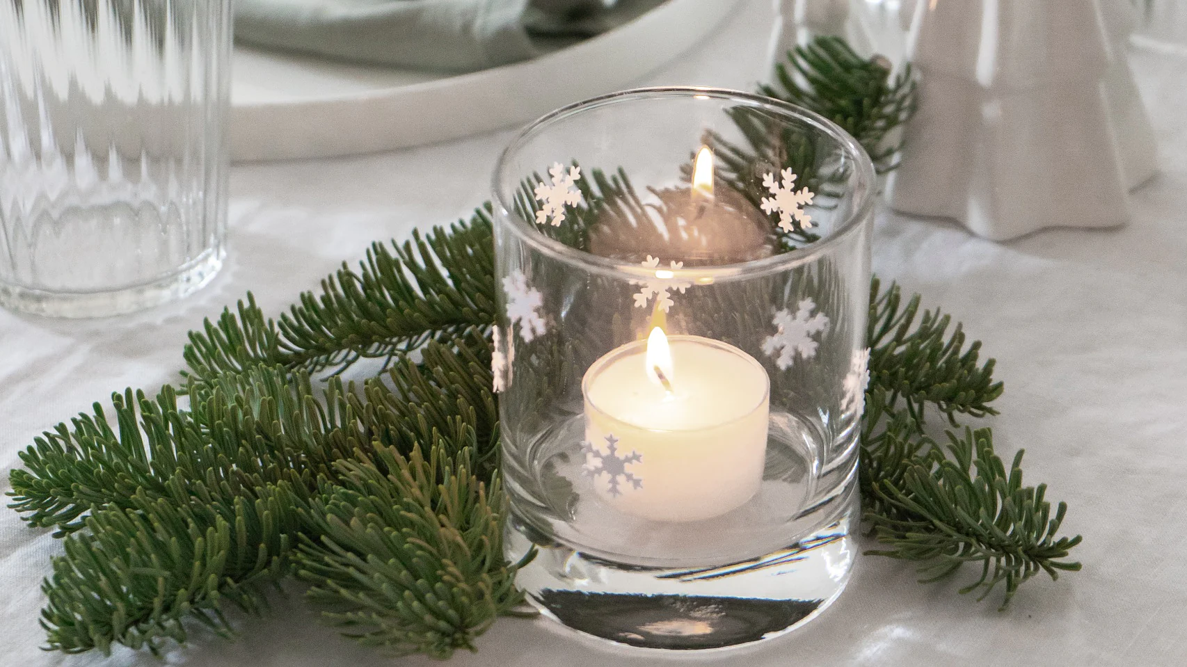 Ein festlich gedeckter Tisch mit einem brennenden Teelicht in einem Glashalter mit Schneeflockenmotiven. Um die Kerze herum sind immergrüne Zweige. Daneben liegen baumförmige Keramikornamente und eine aufgerollte grüne Serviette auf einem Teller, zusammengehalten mit Tesaband. (This text has been generated by AI)