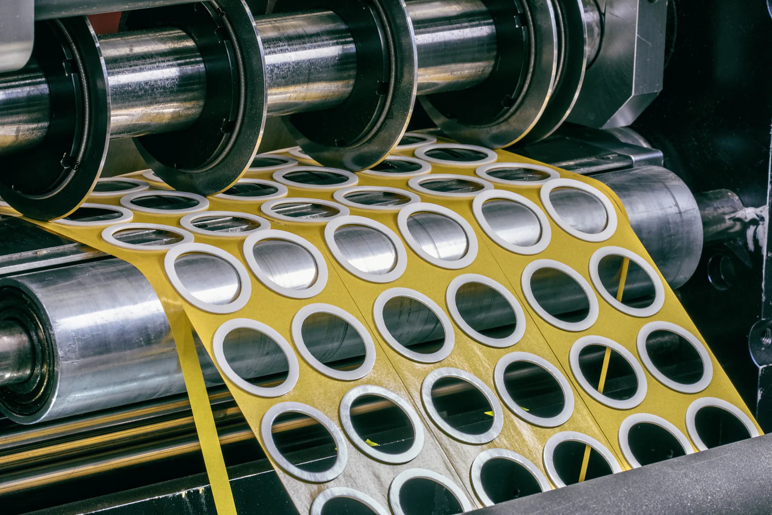 Eine Maschine verarbeitet einen Bogen Klebeetiketten, schneidet sie ab und rollt sie auf eine Spule. Die Etiketten sind rund, mit einer weißen Mitte und einem gelben Rand. Die Maschine verfügt über Metallrollen und komplizierte bewegliche Teile, die mit Tesa-Klebeband umwickelt sind. (This text has been generated by AI)