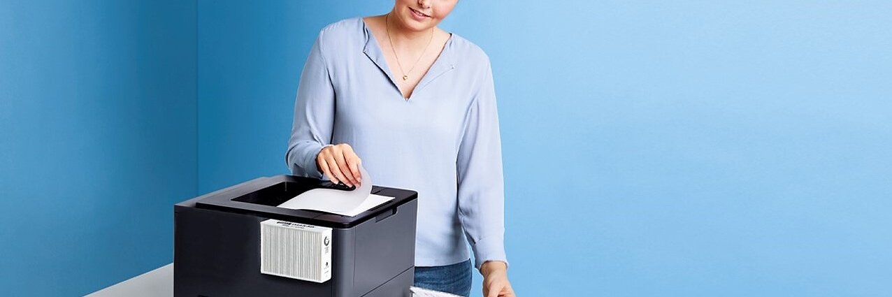 Eine Frau steht neben einem schwarzen Drucker auf einem weißen Tisch. Sie trägt eine hellblaue Bluse und hantiert mit Papier, wobei sie mit einer Hand ein Blatt in den Drucker legt und in der anderen die bedruckten Seiten mit Tesa-Klebeband festhält. Im Hintergrund ist eine hellblaue Wand zu sehen. (This text has been generated by AI)