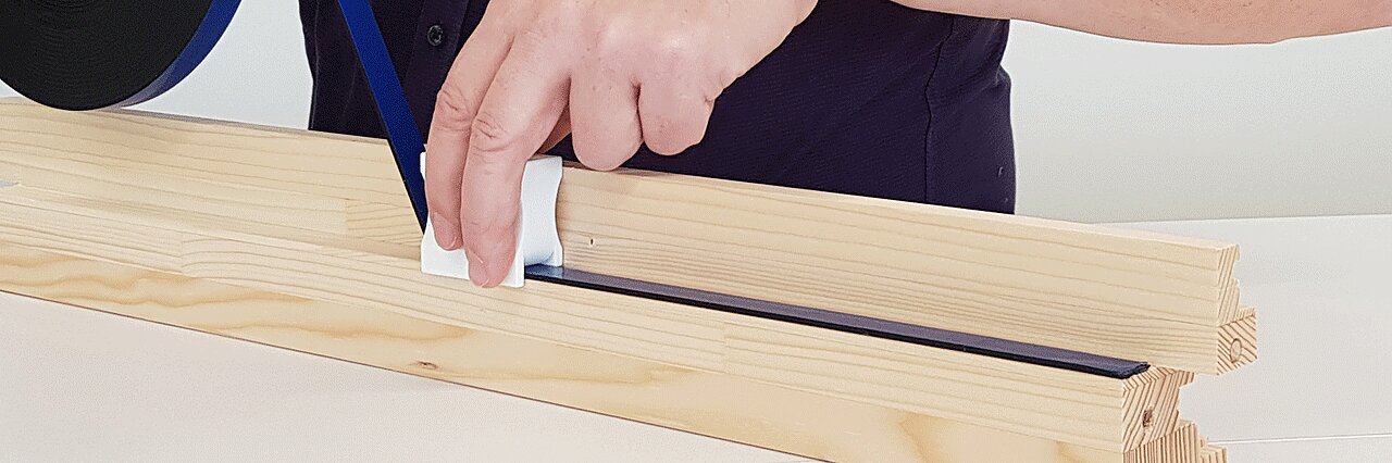 Eine Hand klebt mit einem kleinen weißen Werkzeug einen Streifen schwarzes Tesaband auf einen Holzrahmen. Das Holz hat Rillen, die Person trägt ein dunkles Hemd. Links ist eine Rolle Tesaband teilweise zu sehen. (This text has been generated by AI)