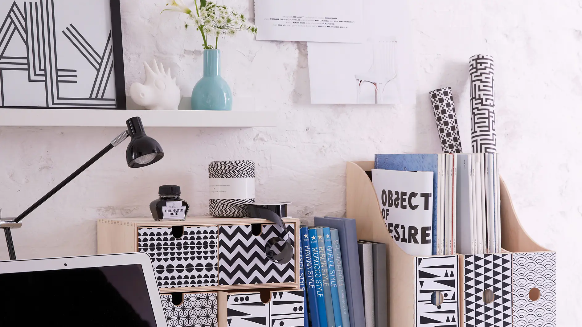 A minimalist desk setup features a laptop, patterned boxes, and a lamp on a white surface. Books and decorative items are arranged neatly. The background is a white brick wall with posters and a small shelf displaying a vase and framed art, all perfectly held in place using tesa tape.