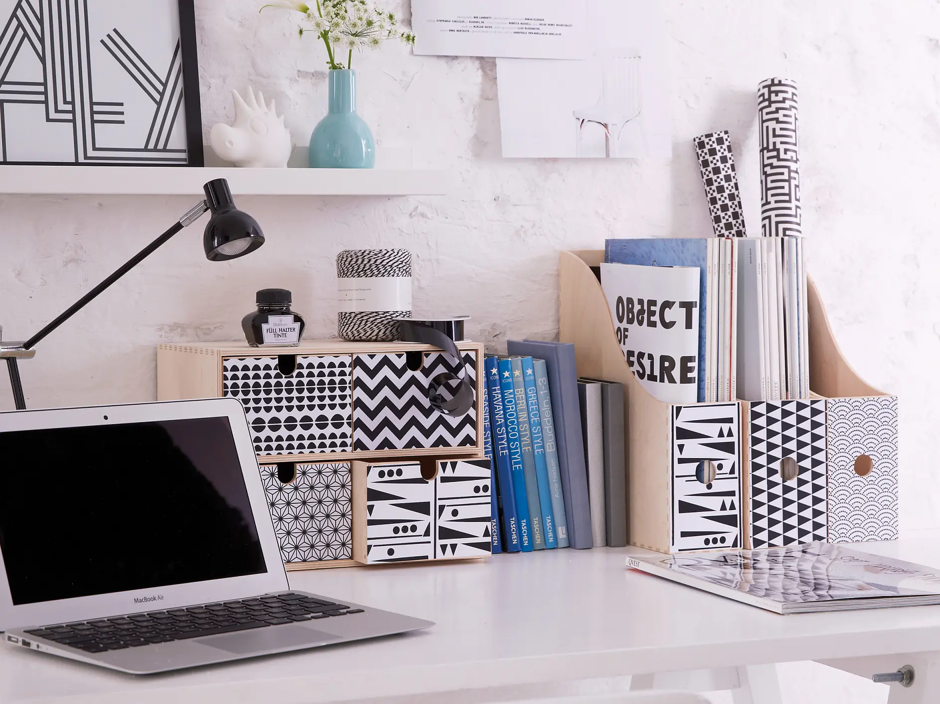 A minimalist desk setup features a laptop, patterned boxes, and a lamp on a white surface. Books and decorative items are arranged neatly. The background is a white brick wall with posters and a small shelf displaying a vase and framed art, all perfectly held in place using tesa tape.