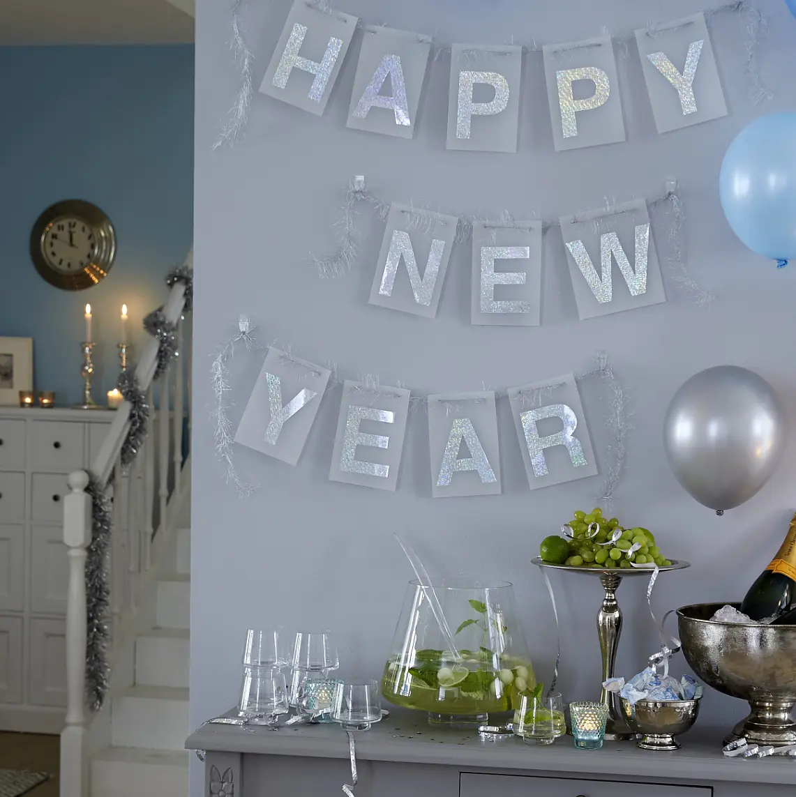 A room decorated for New Years Eve with a Happy New Year banner on the wall, secured using tesa tape. Blue and silver balloons are displayed. A table holds a champagne bucket, glasses, candles, and a bowl of fruits. A white cabinet and staircase are visible in the background. (This text has been generated by AI)