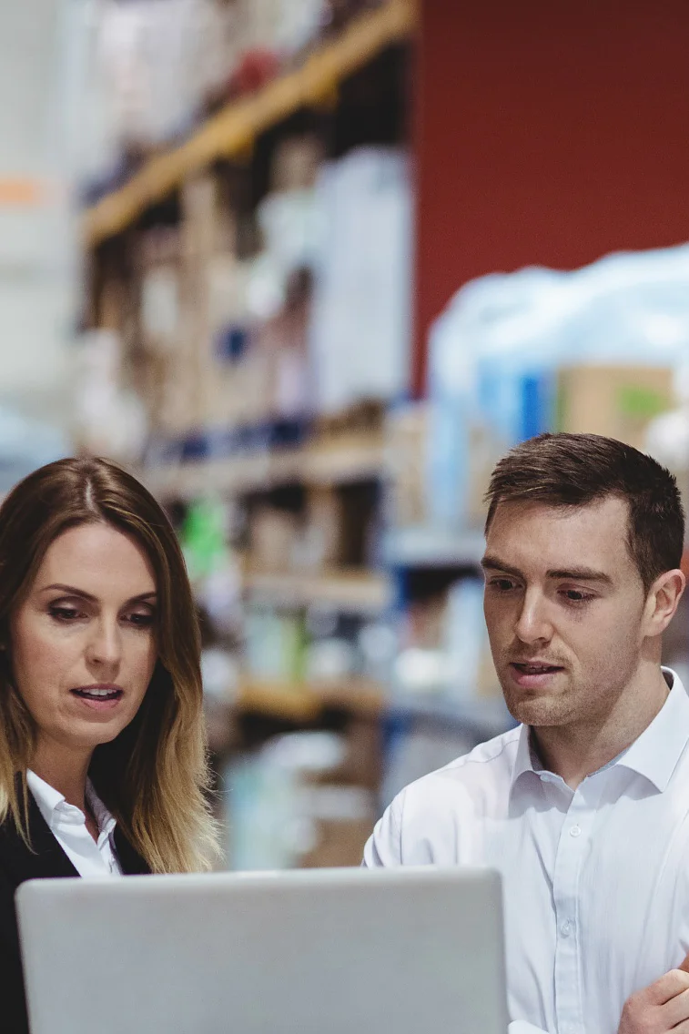 A group of warehouse workers stand by a table filled with packages. Two are engaged with a laptop, while another tallies items using a handheld device. Shelves stocked with boxes and supplies, including tesa tape, are visible in the background. (This text has been generated by AI)