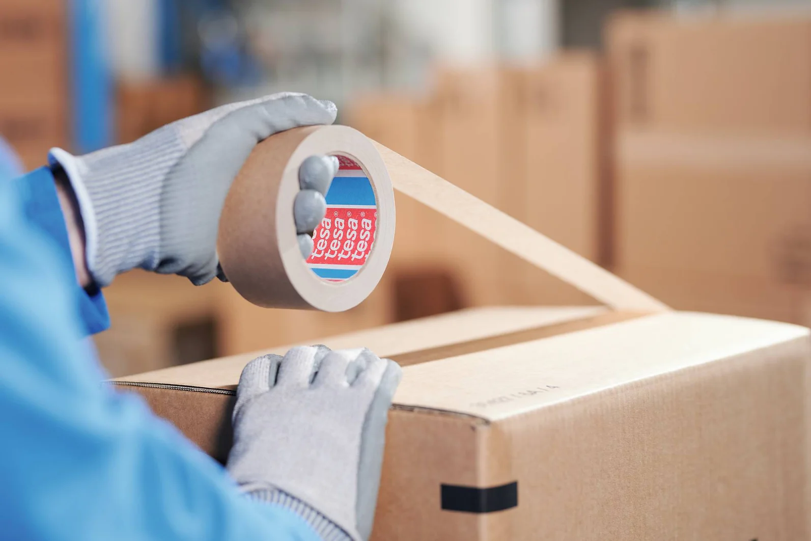 Eine Person mit grauen Handschuhen und blauem Hemd verschließt einen Karton mit beigem Tesa-Verpackungsband. Die Tesa-Bandrolle hat einen roten und einen blauen Kern. Im Hintergrund sind mehrere andere Kartons unscharf zu erkennen. (This text has been generated by AI)