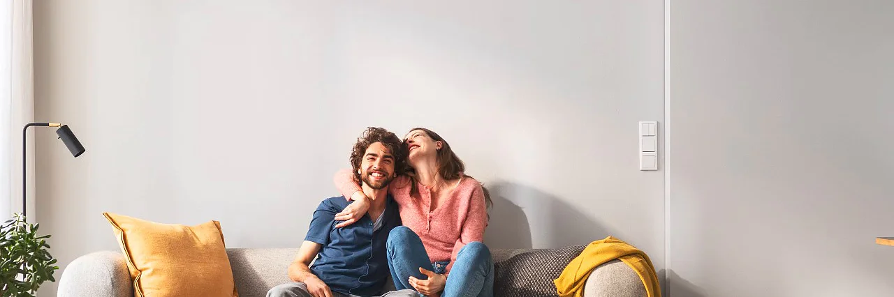 Mit tesa® Bilder aufhängen und glücklich sein Ein Paar sitzt auf dem Sofa. Sie freuen sich, als sich die Bilder wie von selbst aufhängen.