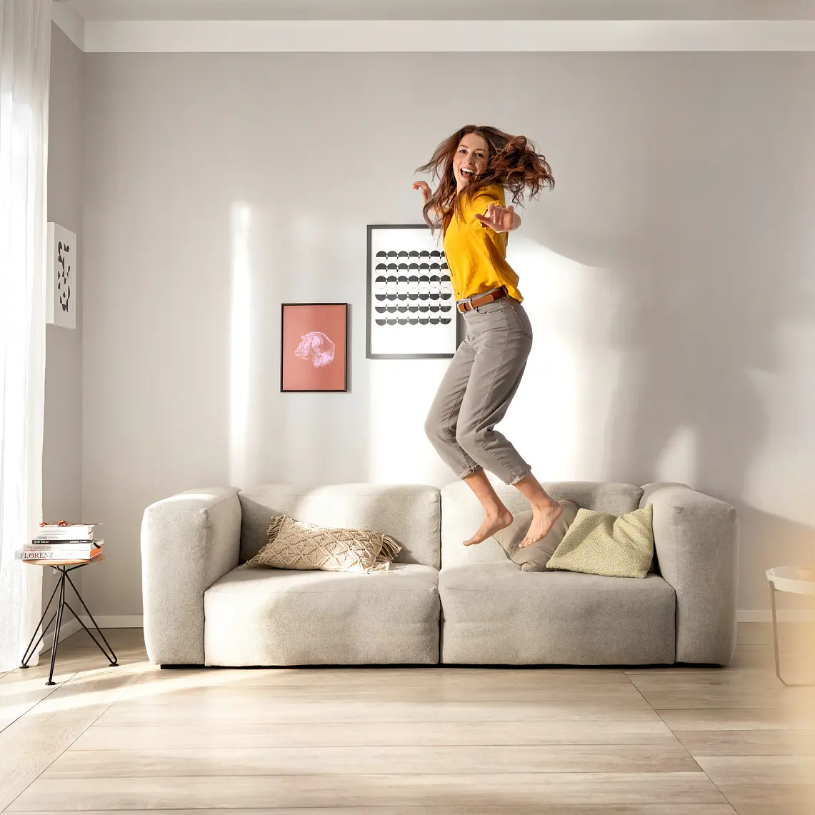 A person with long hair is joyfully jumping on a light gray sofa in a bright room. The person is wearing a yellow shirt and gray pants. The room features light-colored walls, hanging artwork, and large windows with sheer curtains secured using tesa tape. (This text has been generated by AI)