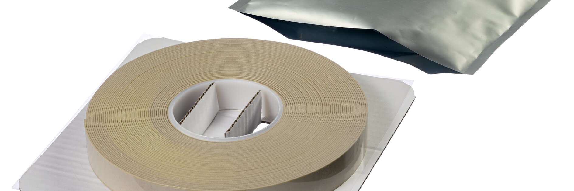 Eine große Rolle beiges Tesa-Klebeband liegt auf einem weißen Kartonhalter. Hinter dem Tesa-Klebeband verbirgt sich eine versiegelte Silberfolienverpackung mit einem rot-weißen Etikett darauf. Beide Artikel sind auf einem schlichten weißen Hintergrund platziert. (This text has been generated by AI)