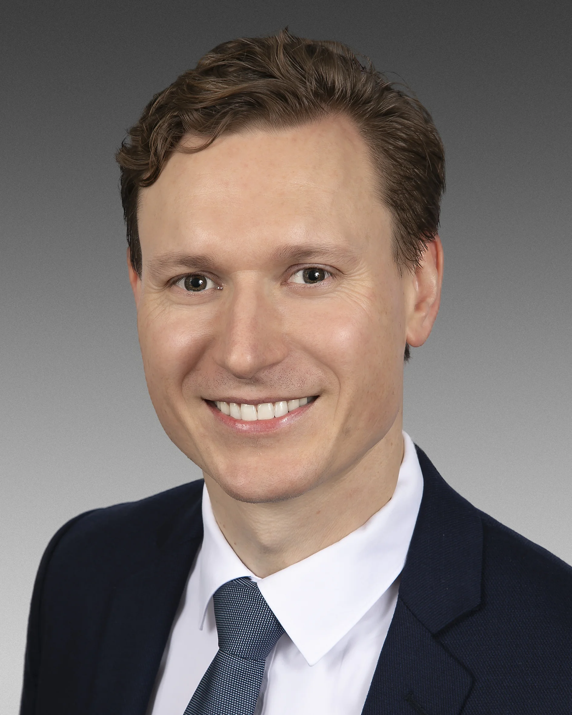 Ein Mann mit kurzen braunen Haaren lächelt auf einem professionellen Porträtfoto. Er trägt einen dunkelblauen Anzug, ein weißes Hemd und eine hellblaue Krawatte. Der Hintergrund ist ein Farbverlauf von Dunkelgrau zu Hellgrau. (This text has been generated by AI)