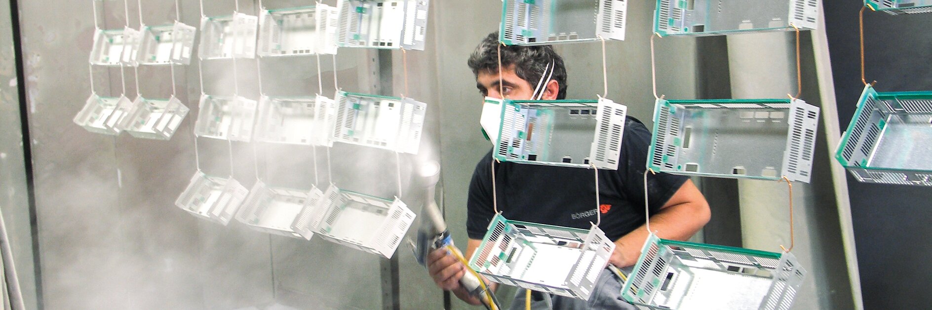 Eine Person mit Mundschutz kniet und trägt mit einer Spritzpistole eine Beschichtung auf mehrere in Reihen hängende Metallrahmen auf. Die Szene spielt in einer Industrieumgebung mit Tesa-Klebebandgittern auf dem Boden. (This text has been generated by AI)