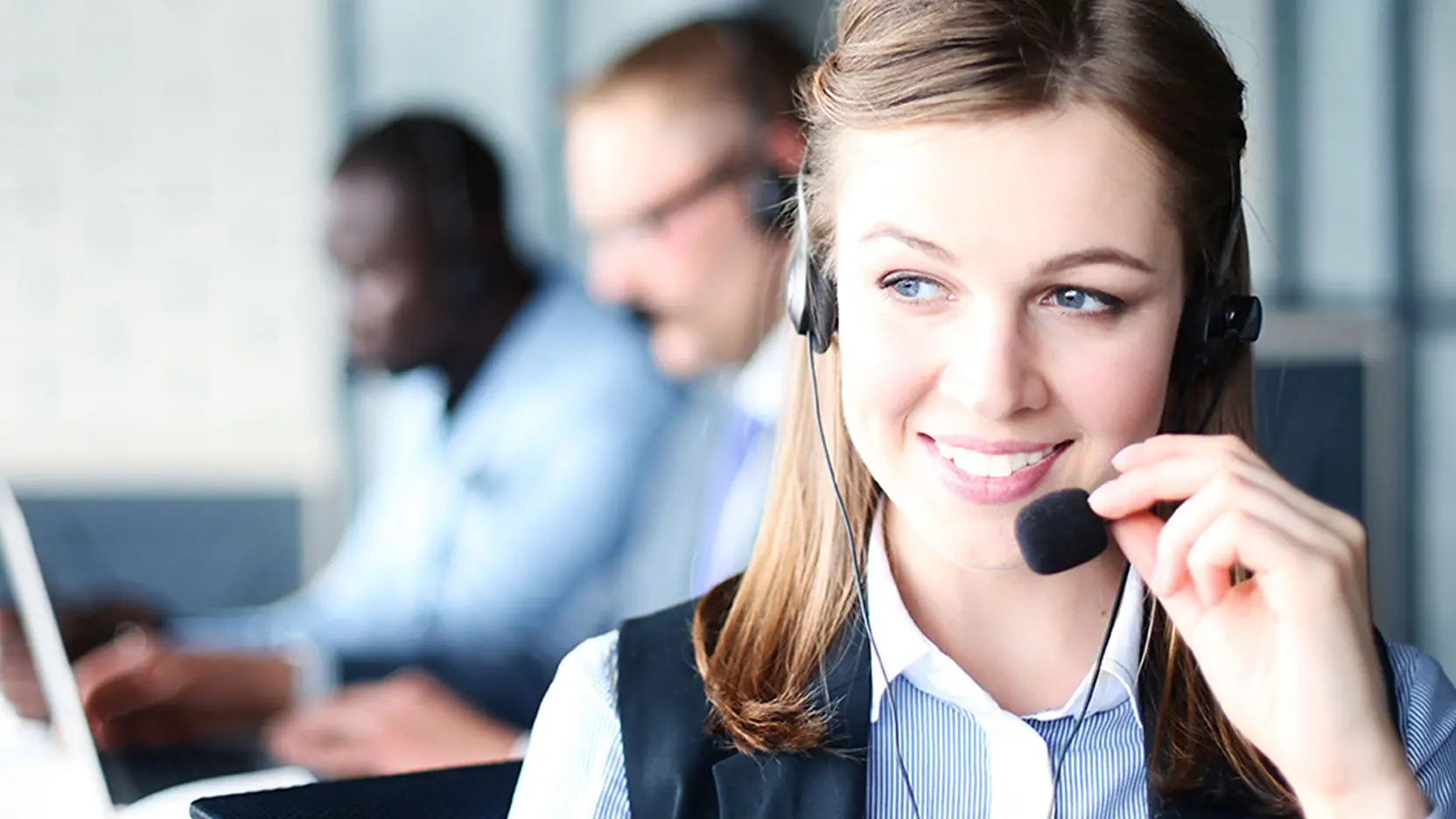 A woman wearing a headset is smiling and looking to the side. She is in focus, while two other people in headsets are in the background, slightly blurred, working on laptops. The setting appears to be an office or a customer service center.