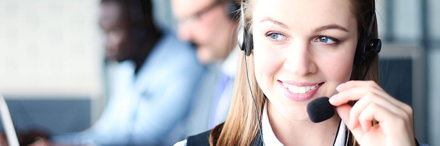 A woman wearing a headset is smiling and looking to the side. She is in focus, while two other people in headsets are in the background, slightly blurred, working on laptops. The setting appears to be an office or a customer service center.