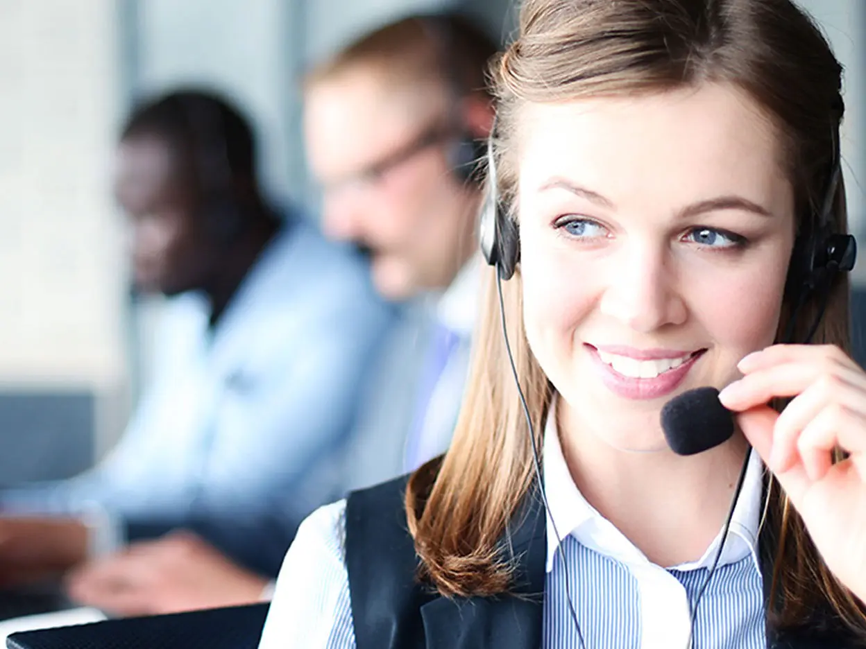 A woman wearing a headset is smiling and looking to the side. She is in focus, while two other people in headsets are in the background, slightly blurred, working on laptops. The setting appears to be an office or a customer service center. (This text has been generated by AI)