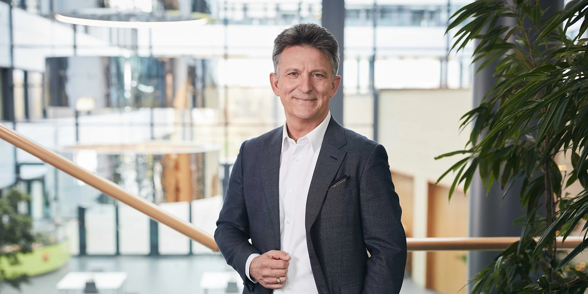 Dr. Norman Goldberg Ein Mann im dunklen Anzug steht drinnen an einem Geländer und lächelt in die Kamera. Im Hintergrund ist moderne Architektur mit großen Fenstern, Pflanzen und weißen Tischen zu sehen, die eine helle und luftige Atmosphäre schaffen. (This text has been generated by AI)