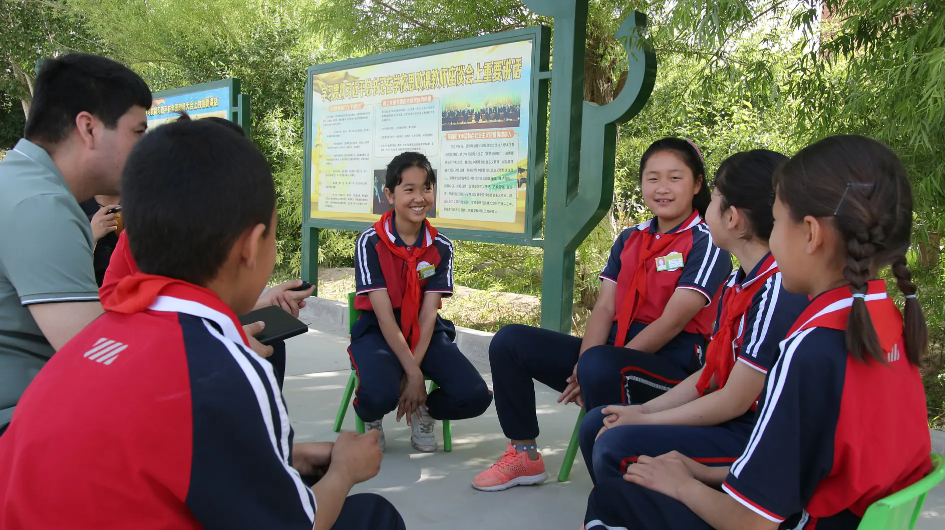 A group of children in red and blue uniforms is seated on small chairs outdoors, engaged in conversation with an adult wearing a green shirt. They are near a large informational sign featuring text and images. Trees are visible in the background. (This text has been generated by AI)