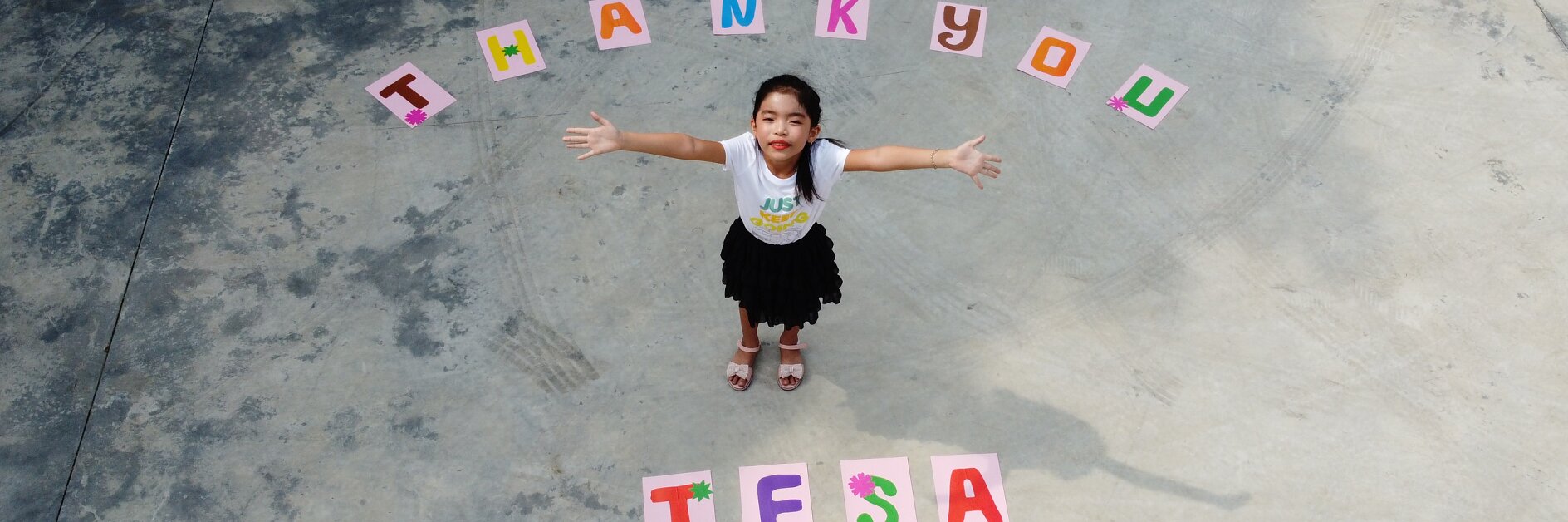 Child standing on concrete with arms outstretched. Above her, colorful letters spell THANK YOU, and below her, tesa tape. The child is wearing a white shirt and black skirt. The scene is shot from above.
