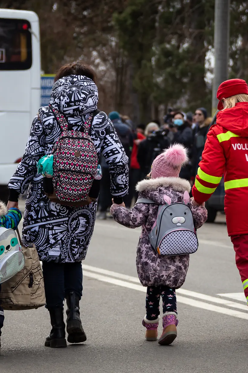 A woman and two children walk down a street near parked vehicles. The children wear backpacks and winter clothing. A volunteer wearing a red uniform assists them, using tesa tape to secure a large white bag. A bus and several people are visible in the background. (This text has been generated by AI)