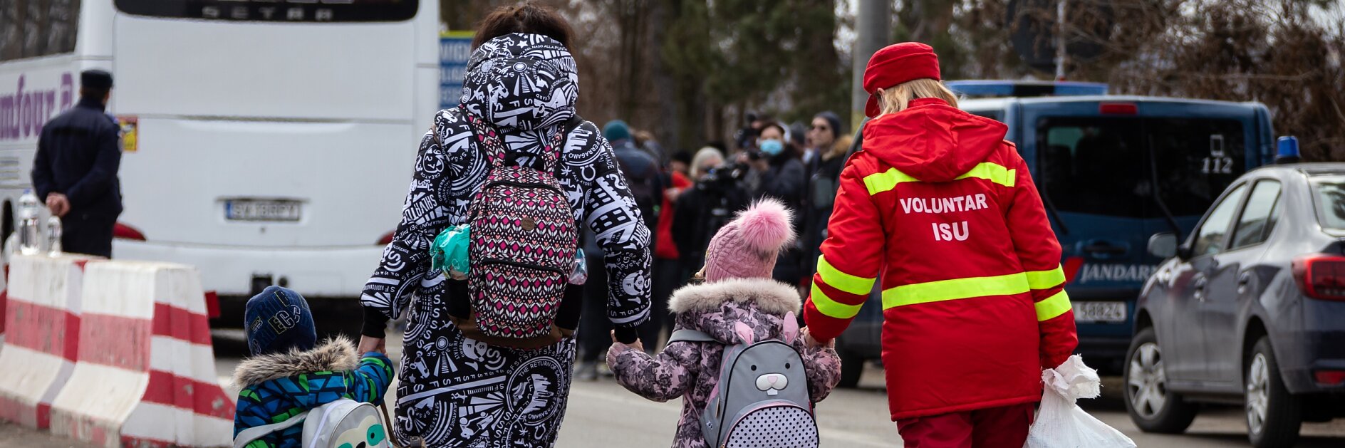 A woman and two children walk down a street near parked vehicles. The children wear backpacks and winter clothing. A volunteer wearing a red uniform assists them, using tesa tape to secure a large white bag. A bus and several people are visible in the background.