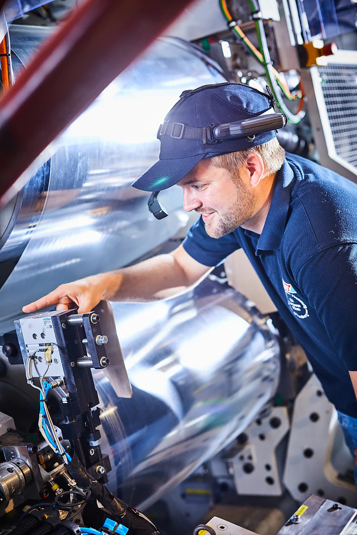 Ein Mann mit Mütze und Poloshirt bedient eine Maschine mit großen zylindrischen Teilen. Mit seiner linken Hand justiert er ein Bauteil. Die Umgebung wirkt industriell, mit sichtbaren Kabeln und Metalloberflächen. Licht wird von der Maschine und nahegelegenen Rollen Tesaband reflektiert. (This text has been generated by AI)