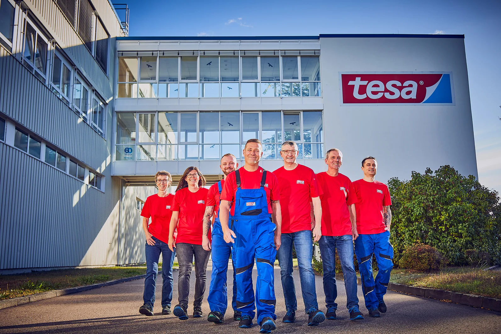 Sieben Menschen in roten Hemden stehen vor einem Gebäude mit einem „Tesa Tape“-Schild. Sie stehen an einem sonnigen Tag im Freien und eine Person in der Mitte trägt einen blauen Overall. Das Gebäude ist weiß und hat mehrere Fenster. (This text has been generated by AI)