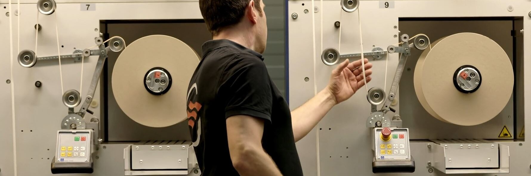 Eine Person in einem schwarzen Hemd bedient eine Maschine, an der große Rollen Tesa-Klebeband beteiligt sind. Die Maschine hat Bedienfelder mit Knöpfen und Drehreglern. Die Szene scheint in einer Industrie- oder Fertigungsumgebung zu sein. (This text has been generated by AI)