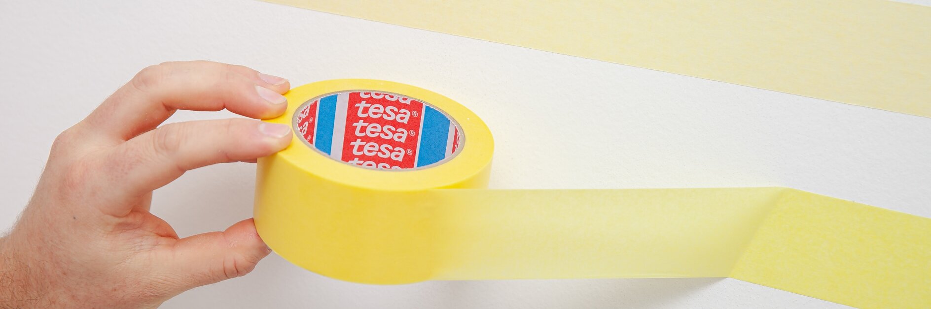 Eine Hand hält eine Rolle gelbes Tesaband vor eine weiße Wand. Ein Streifen Tesaband wird abgerollt. Auf der Innenseite ist das Etikett der Tesabandrolle zu sehen, das ein rot-blaues Motiv mit dem Wort „Tesa“ zeigt. (This text has been generated by AI)