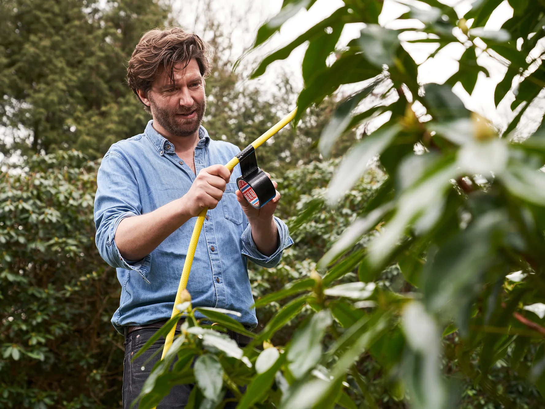 Ein Mann in Jeanshemd inspiziert einen gelben Gartenschlauch im Grünen. Er scheint sich darauf zu konzentrieren, eine Armatur mit Tesa-Klebeband am Schlauch zu befestigen oder einzustellen. Im Hintergrund sind üppige grüne Büsche zu sehen. (This text has been generated by AI)