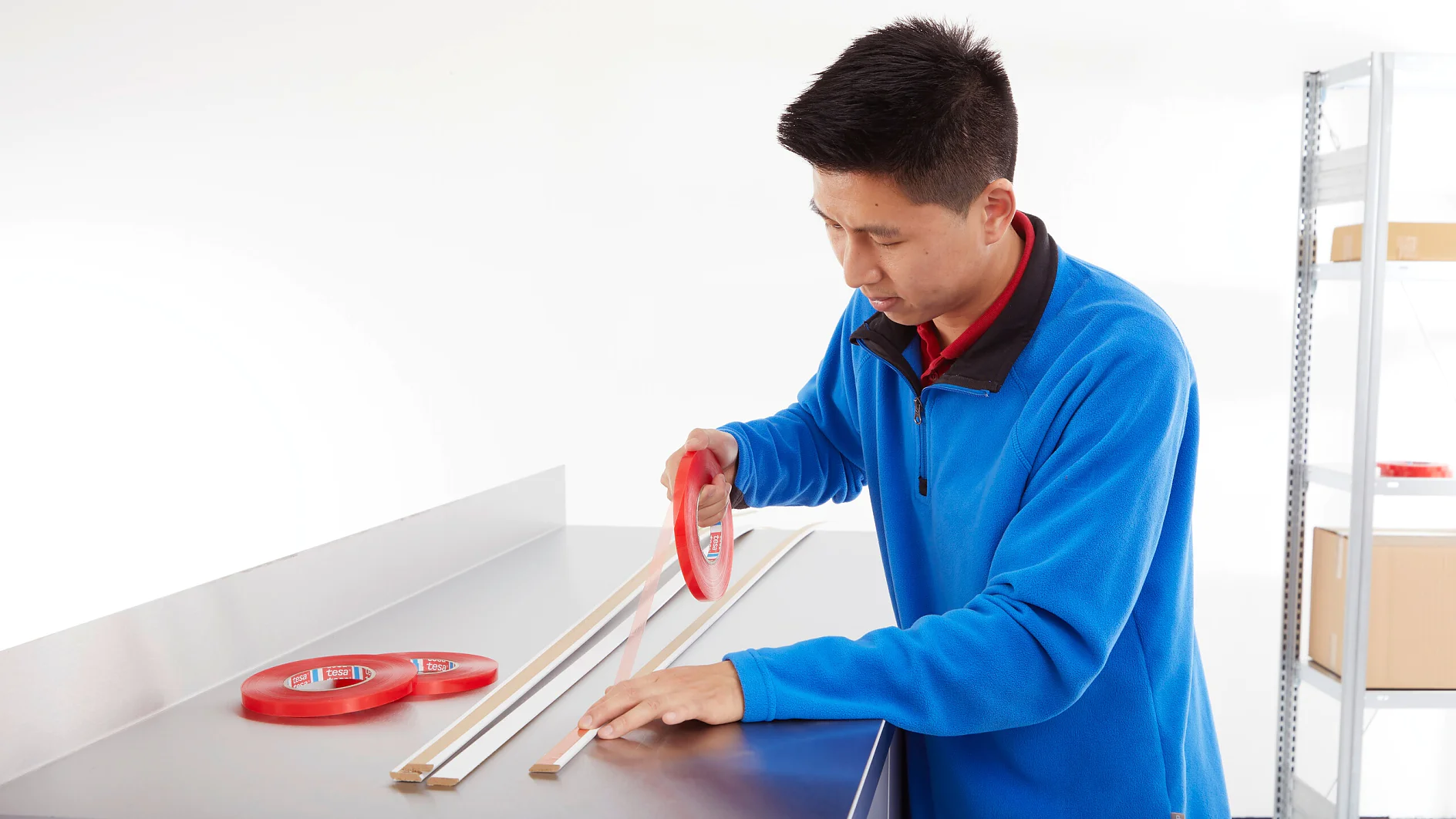 Eine Person in einer blauen Jacke klebt doppelseitiges Tesa-Klebeband auf Holzleisten auf einem Metalltisch. Daneben liegen mehrere rote Tesa-Klebebandrollen. Der Hintergrund ist weiß, rechts sind teilweise Regale zu sehen. (This text has been generated by AI)