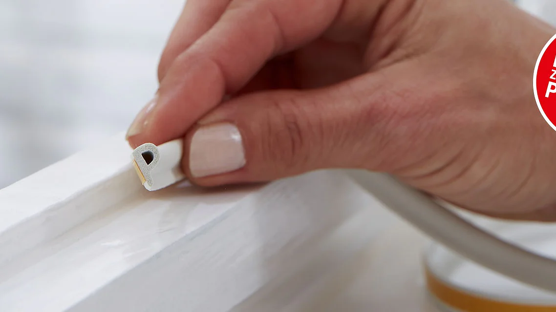 A person is applying self-adhesive weatherstripping to a window frame. A roll of tesa tape weatherstripping is visible beside the frame. Theres a red circle with text in German promoting a product. (This text has been generated by AI)