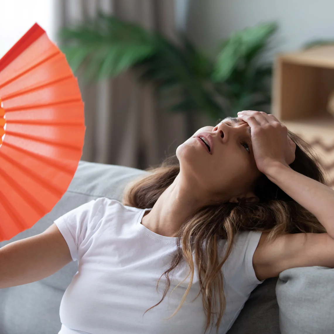 Overheated woman sitting on couch, waving orange paper fan close up, girl feeling unwell, suffering from heating at home, feeling discomfort, hot summer weather or fever, sitting on couch alone