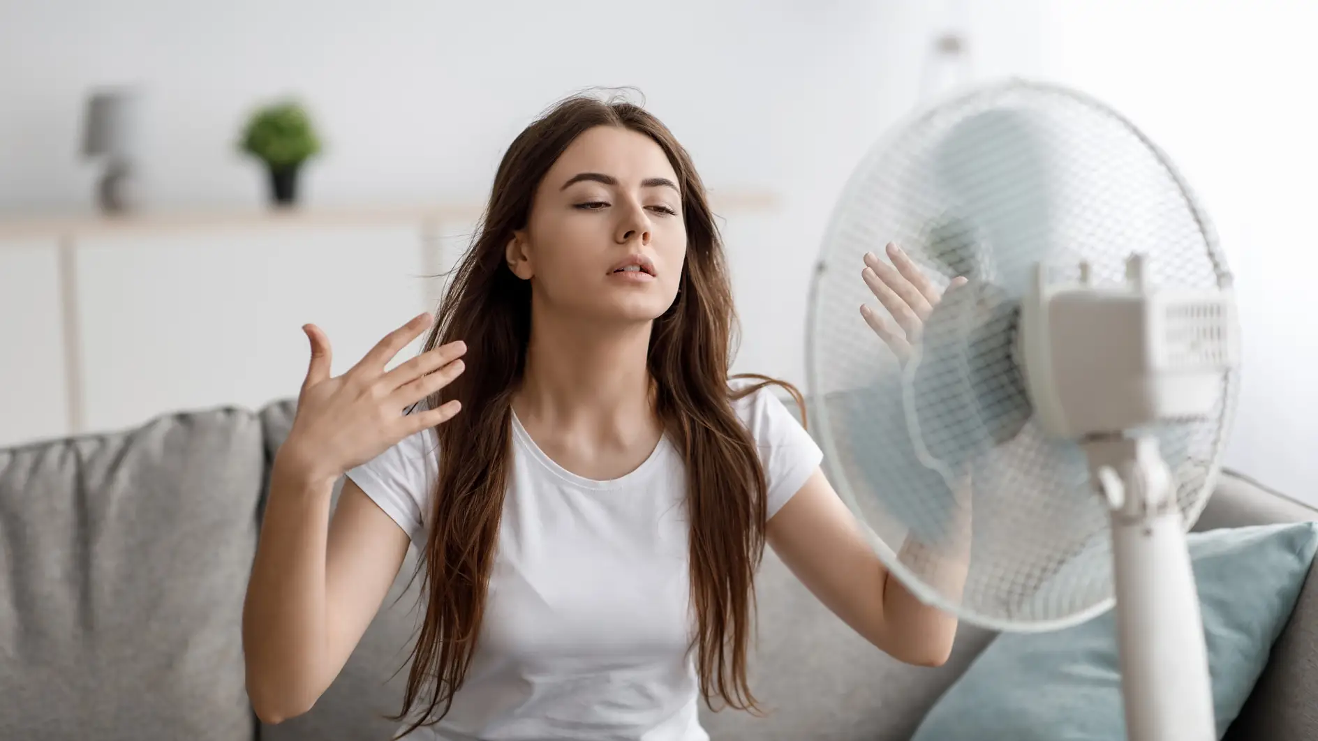 Sad young european woman suffers from unbearably too hot weather, catches cold air from fan in living room interior. Home without air conditioning, lady tired from summer heat, waves arms to cool down