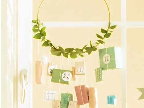 An einem goldenen Ring, der mit grünen Blättern verziert ist, hängt ein handgefertigter Adventskalender. An dem Ring hängen verschiedene kleine, verpackte Päckchen, die jeweils mit einer Nummer zwischen 1 und 24 beschriftet sind, vor einem hellen Hintergrund. (This text has been generated by AI)