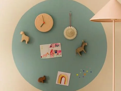 An einer Wand hängt ein großer blaugrüner Kreis mit Gegenständen wie einer kleinen Uhr, Dekoartikeln und Kunstwerken, die mit Tesa-Klebeband befestigt sind. Es gibt Tierfiguren, eine Postkarte und eine Kinderzeichnung eines Regenbogens. Rechts ist ein Teil einer beigen Stehlampe zu sehen. (This text has been generated by AI)