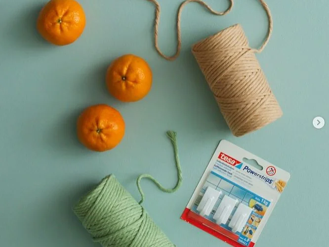 Auf einem hellblauen Untergrund sind drei Orangen, zwei Rollen Kordel (eine beige, eine grün) und eine Packung Tesafilm-Wandhaken angeordnet. (This text has been generated by AI)