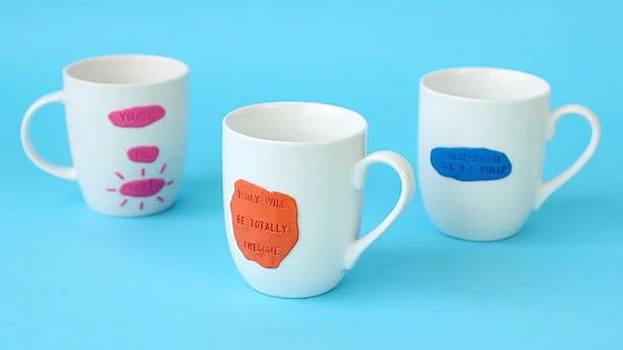 Drei weiße Tassen auf blauem Hintergrund, jede mit bunten Tondekorationen geschmückt. Die vordere Tasse ist mit einem orangefarbenen Schild mit einem mit Tesaband befestigten Text versehen, die linke Rückseite mit einem rosa Schild mit einem mit Tesaband befestigten Text und die rechte Rückseite mit einem blauen Schild mit einem ebenfalls mit Tesaband befestigten Text. (This text has been generated by AI)