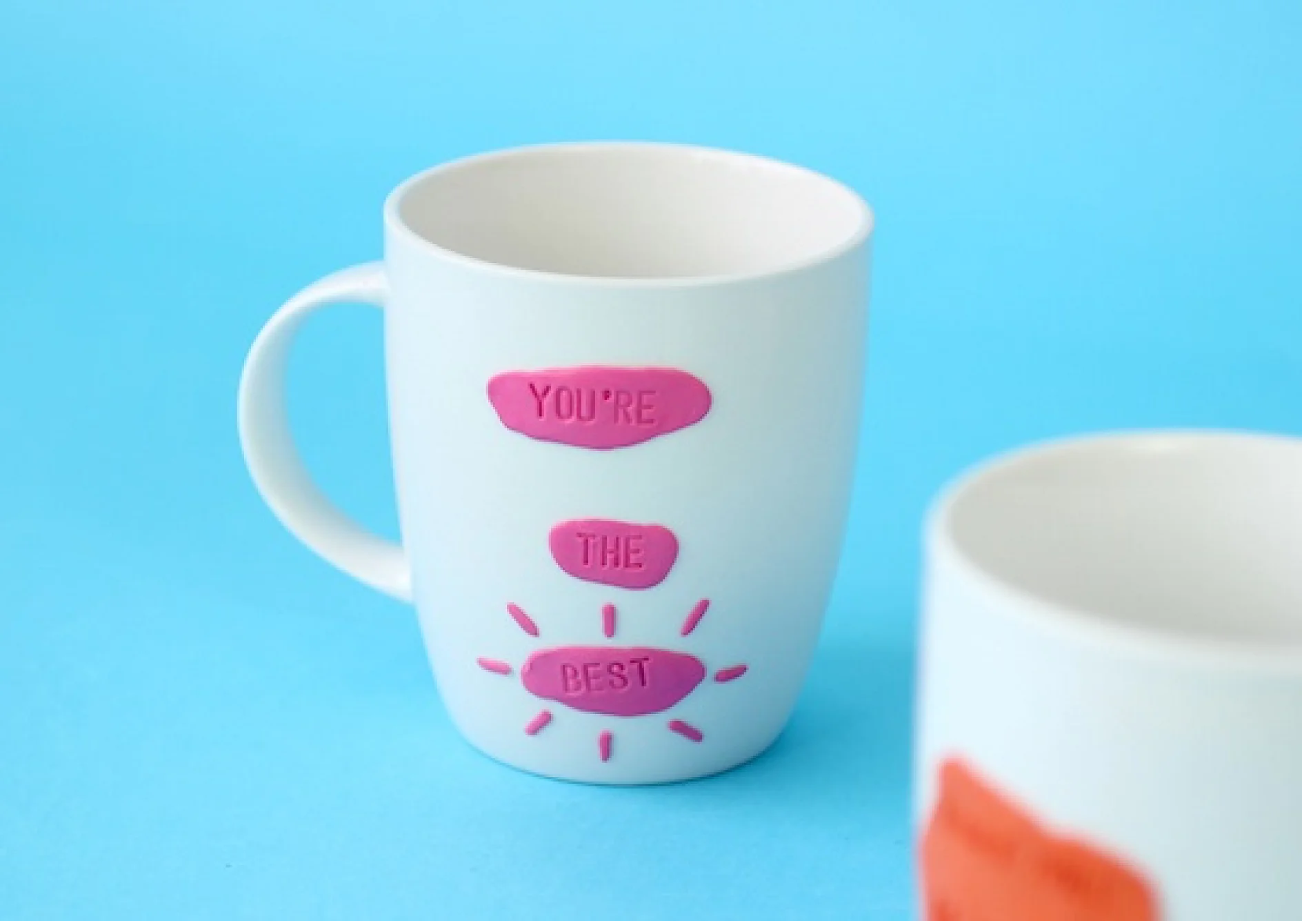 Eine weiße Tasse ist mit rosa Buchstaben verziert, auf denen vertikal „DU BIST DER BESTE“ steht. Weitere rosa Linien bilden unterhalb des Textes einen Sonnenstrahl. Eine weitere Tasse mit orangefarbenem Motiv, die mit Tesa-Klebeband befestigt ist, ist teilweise im Vordergrund zu sehen. Der Hintergrund ist blau. (This text has been generated by AI)