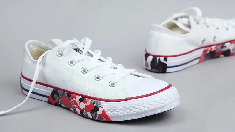 Weiße Low-Top-Sneaker sind vor einem einfarbigen Hintergrund zu sehen. Die Schuhe haben rote und schwarze abstrakte Farbspritzer an den Seiten und einen roten Streifen entlang der Sohle. Bei einem Schuh sind die Schnürsenkel offen, der andere ist weiter hinten positioniert und mit Tesa-Klebeband befestigt. (This text has been generated by AI)