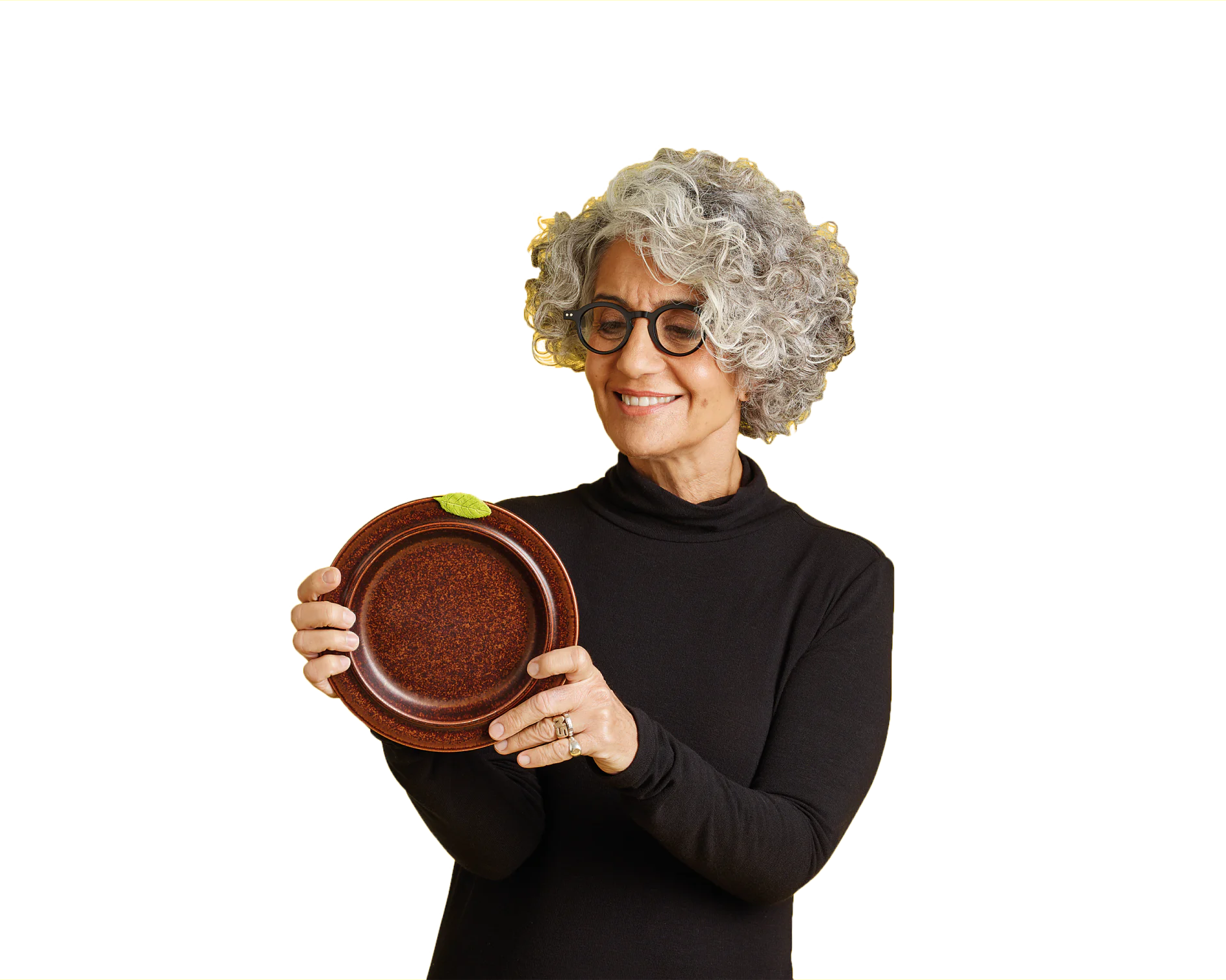 Silvina-posing-with-plate-1-CMYK Eine Person mit lockigem grauem Haar und Brille lächelt und hält eine braune Keramikschale mit einem kleinen grünen Tesa-Klebeband-Etikett in der Hand. Die Person trägt einen schwarzen Rollkragenpullover. Der Hintergrund ist schlicht. (This text has been generated by AI)