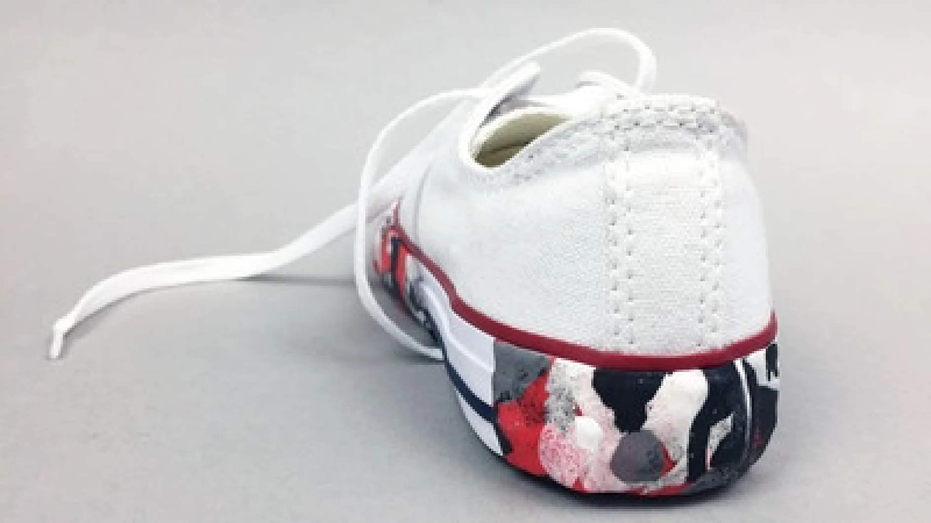 Ein weißer Sneaker mit roten und schwarzen Farbspritzern auf der Sohle, von hinten betrachtet. Der Schuh ist mit weißen Schnürsenkeln befestigt, der Hintergrund ist hellgrau. (This text has been generated by AI)