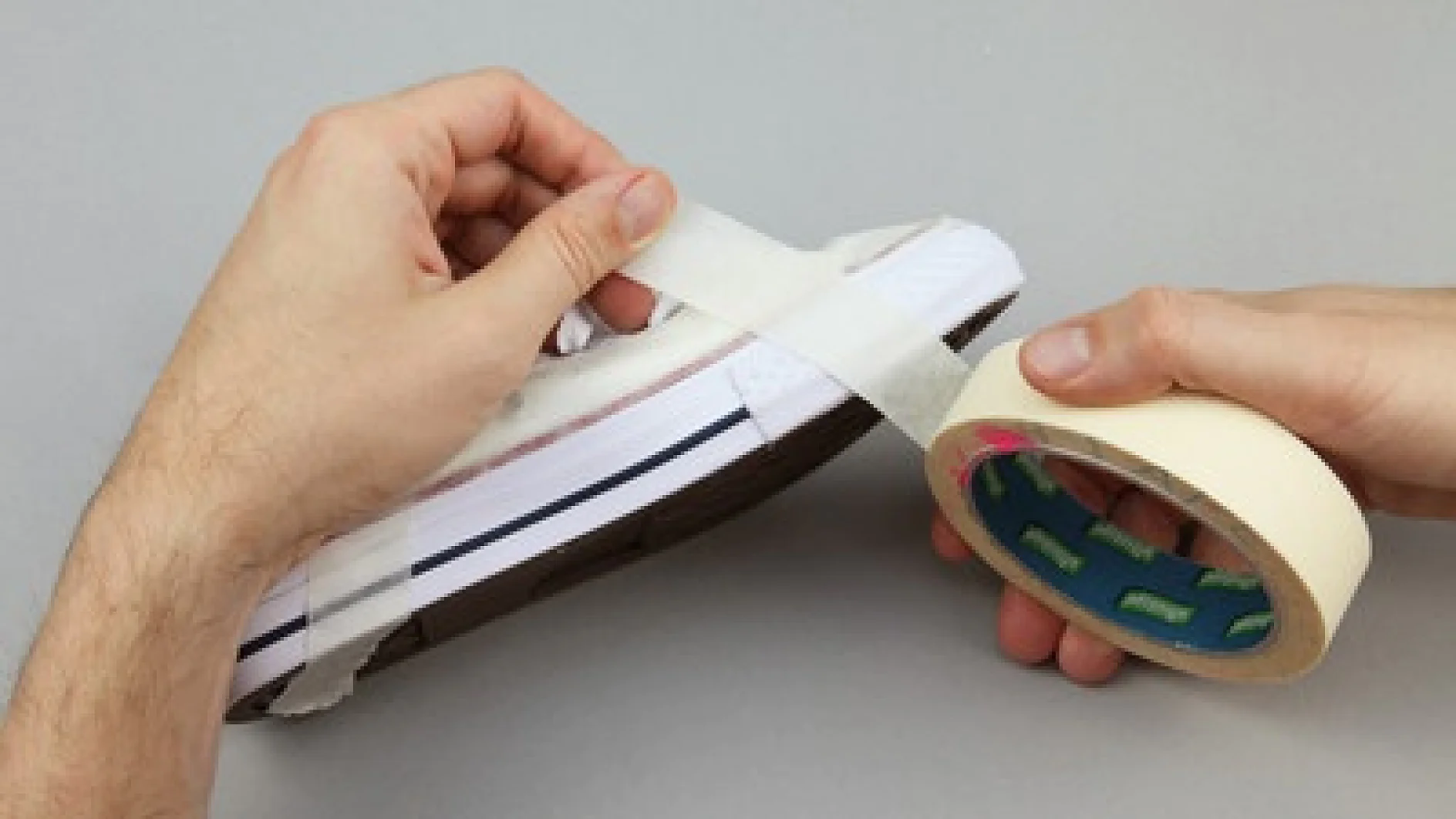 Eine Person klebt Tesa-Klebeband an die Seite eines weißen Sneakers mit brauner Sohle. Eine Hand hält den Sneaker, während die andere Tesa-Klebeband von einer Rolle abzieht. Der Hintergrund ist schlicht grau. (This text has been generated by AI)