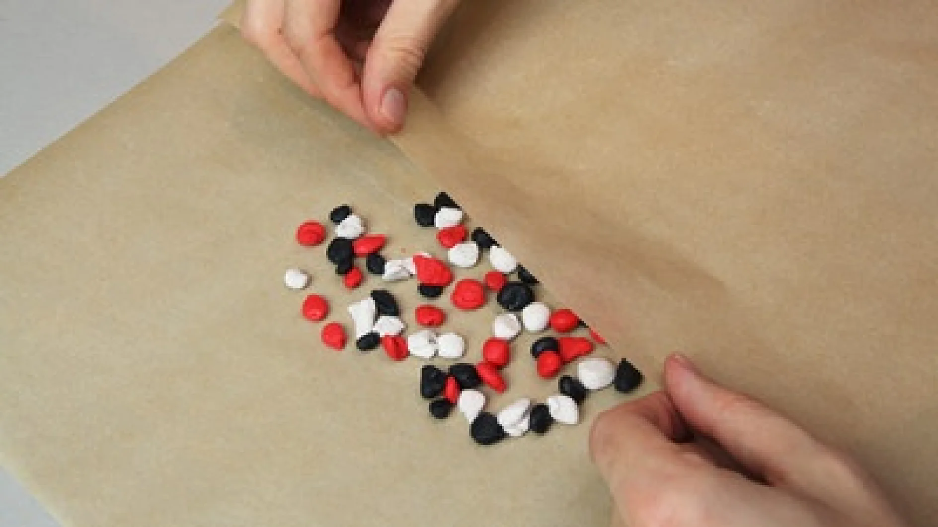 Hände falten ein Blatt braunes Wachspapier über kleine schwarze, rote und weiße Perlen, die auf einer flachen Oberfläche angeordnet und mit Tesaband befestigt sind. (This text has been generated by AI)