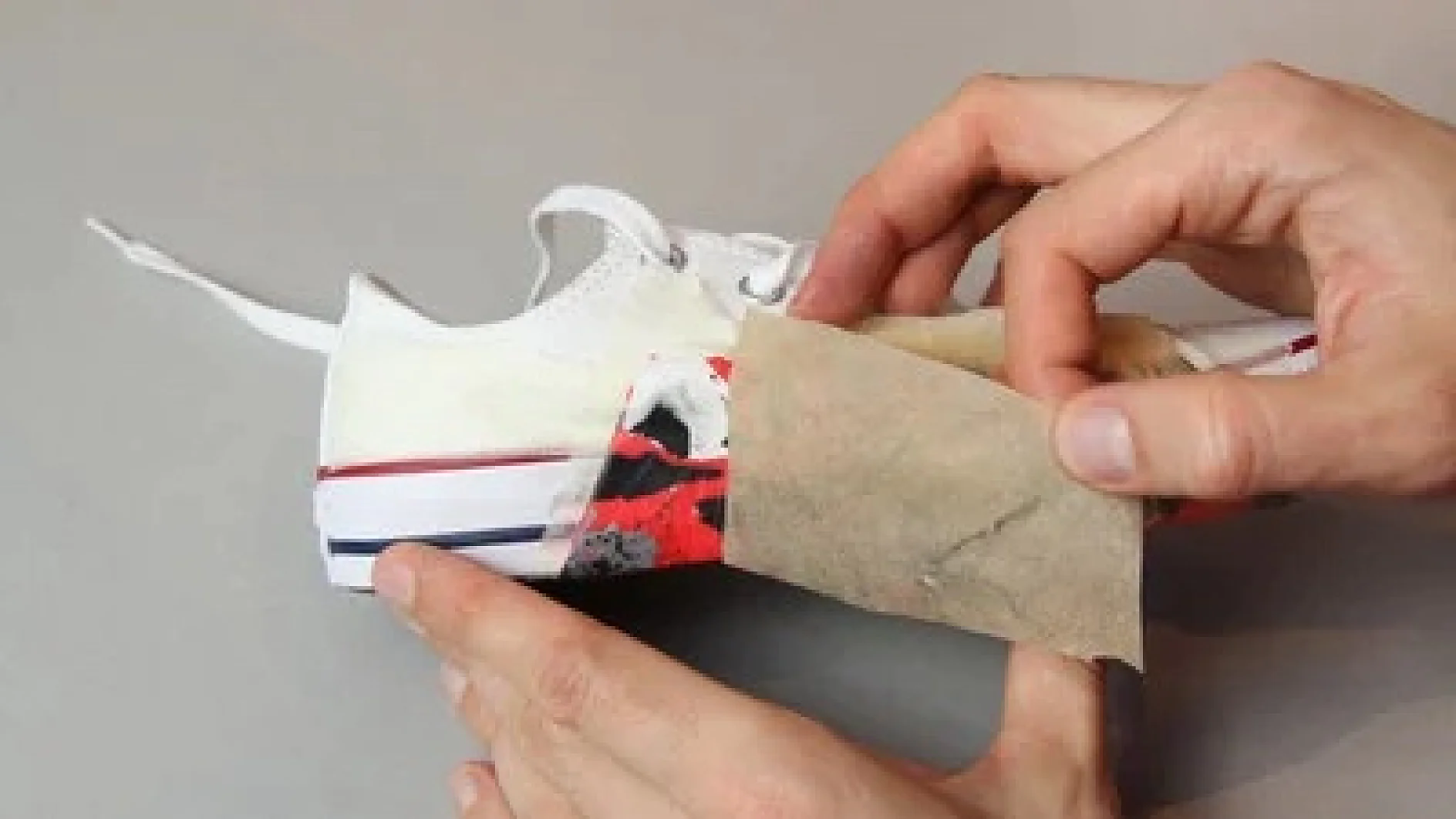 Es sind Hände zu sehen, die ein Stück Schleifpapier an der Seite eines weißen Schuhs mit roten und schwarzen Farbflecken anbringen. Der Schuh hat weiße Schnürsenkel und eine sichtbare Sohle mit roten und blauen Streifen. Die Kanten sind mit Tesa-Klebeband befestigt, damit beim Schleifen Präzision gewährleistet ist. Der Hintergrund ist schlicht grau. (This text has been generated by AI)