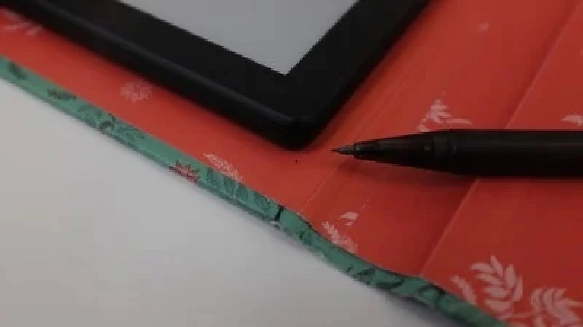 Ein schwarzer Stift liegt auf einem rot-grün gemusterten Stoff. Der Stift befindet sich in der Nähe der Ecke eines schwarzen Tablet-Bildschirms. Der Hintergrund ist weiß und der Tablet-Bildschirm wurde mit Tesa-Klebeband befestigt. (This text has been generated by AI)