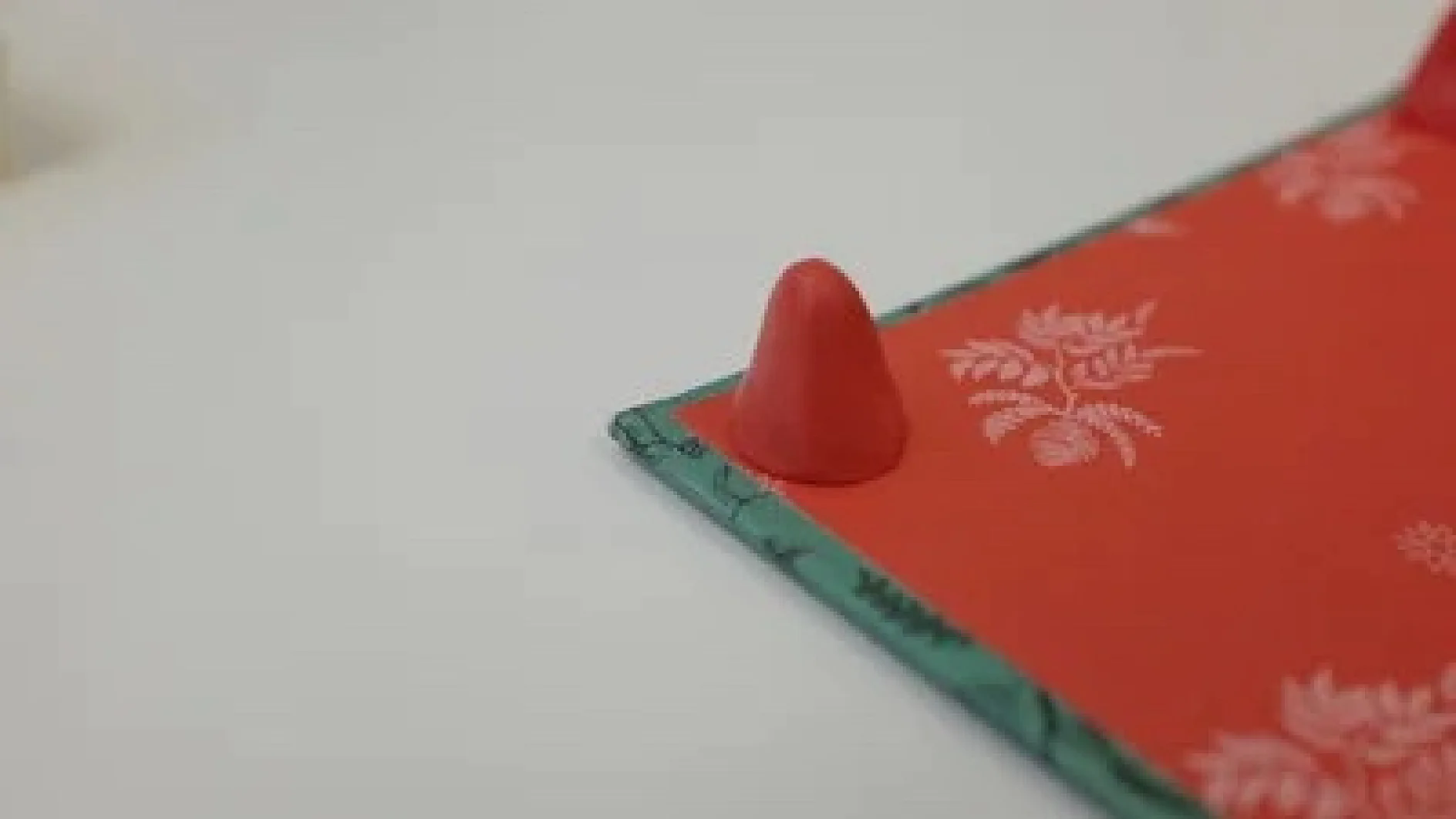 Ein kleines, rotes, kegelförmiges Objekt wird auf einer flachen Oberfläche neben einem teilweise sichtbaren Buch oder Ordner mit einem rot-grünen Einband mit Blumenmuster platziert. Die Oberfläche scheint weiß zu sein, daneben befindet sich etwas Tesa-Klebeband. (This text has been generated by AI)