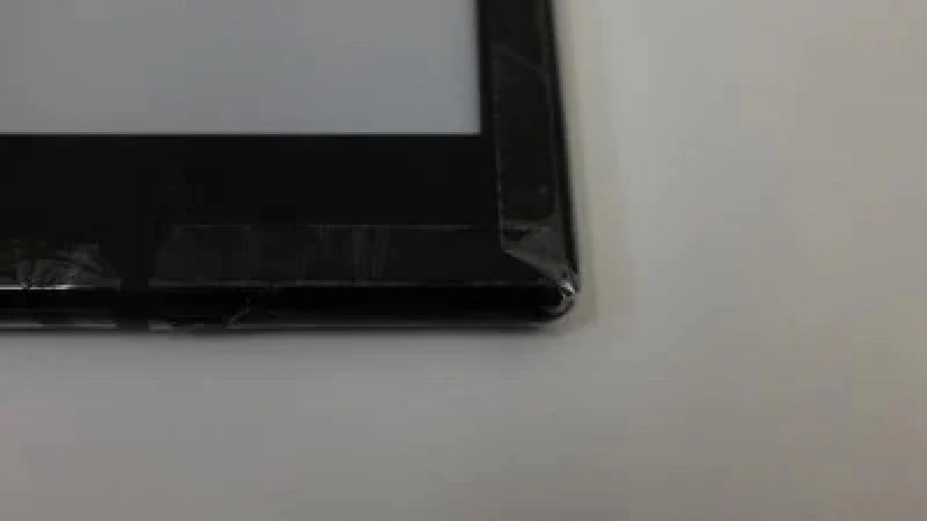 Das Bild zeigt die Ecke eines schwarzen elektronischen Geräts, möglicherweise eines Tablets oder E-Readers, das auf einer weißen Oberfläche liegt. Der Bildschirm des Geräts ist teilweise sichtbar und an der Kante ist durchsichtiges Tesa-Klebeband angebracht, was auf eine kleine Reparatur oder einen Schutz hindeutet. (This text has been generated by AI)