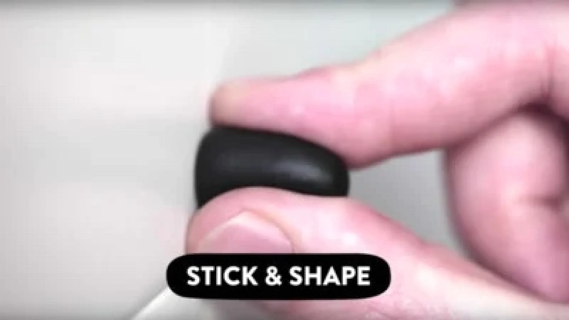 Eine Hand hält ein kleines, schwarzes, rundes Objekt. Der Hintergrund ist unscharf. Auf der Textbeschriftung am unteren Rand steht „Tesa Tape STICK & SHAPE“. (This text has been generated by AI)
